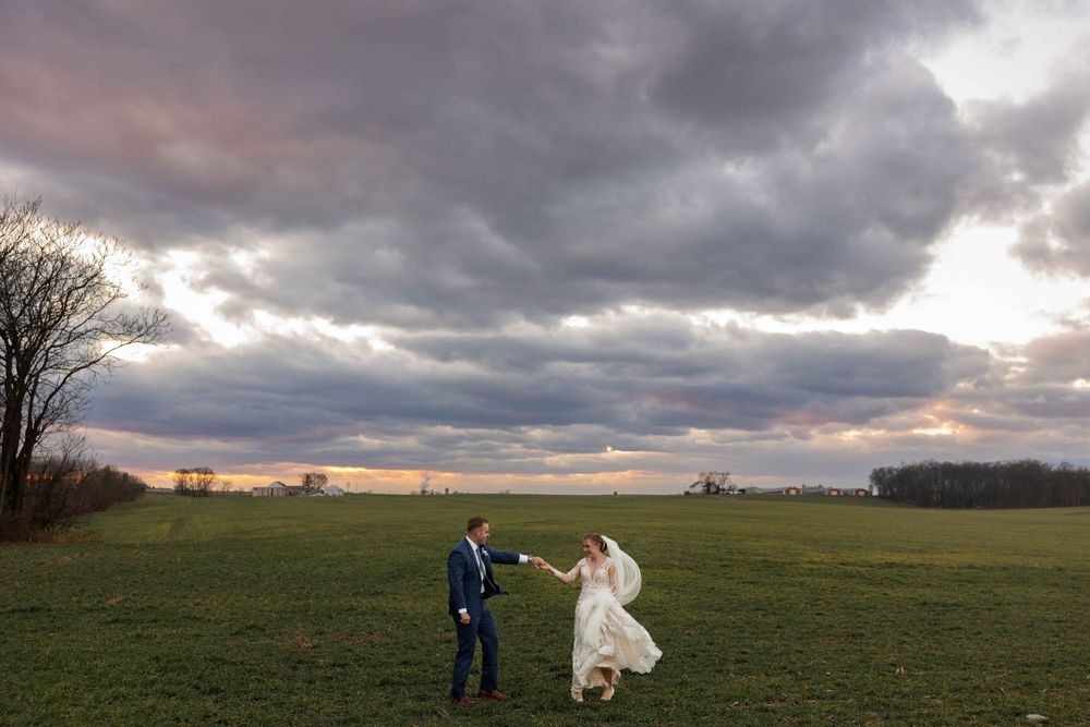 Couple in wedding attire dancing in an open field during a dramatic sunset with stormy clouds.
