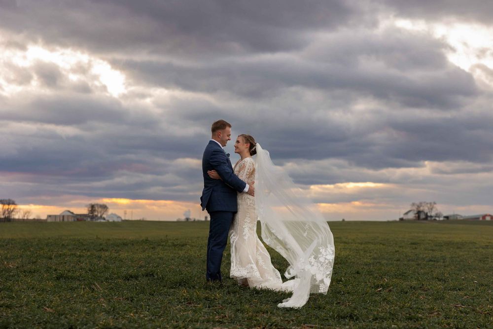 Romantic portrait of newlyweds with flowing veil against dramatic sunset sky in open field.
