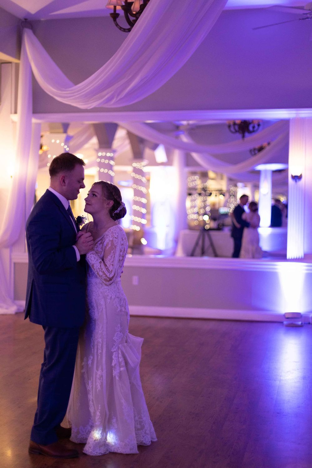 Couple sharing their first dance in a purple-lit reception venue with elegant white draping.