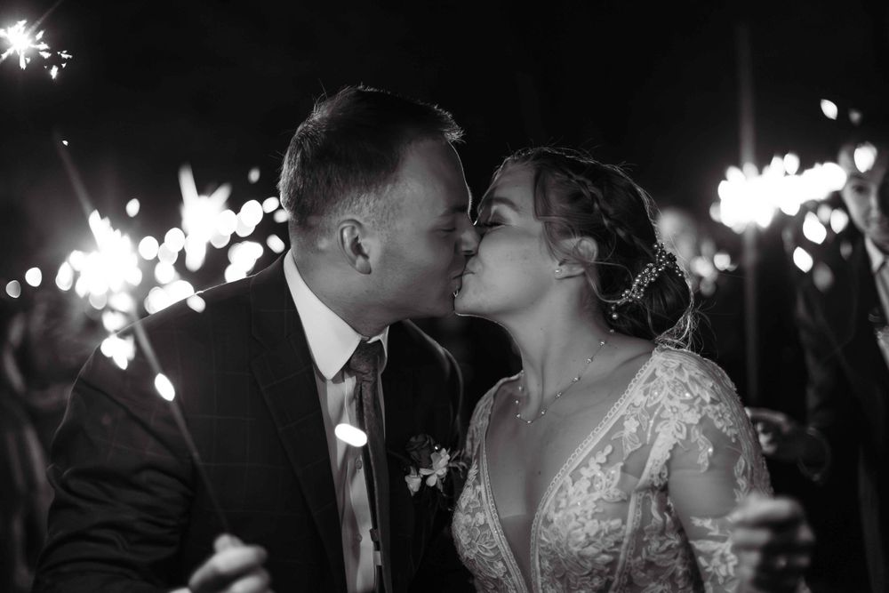 Black and white photo of newlyweds kissing surrounded by sparklers at night.
