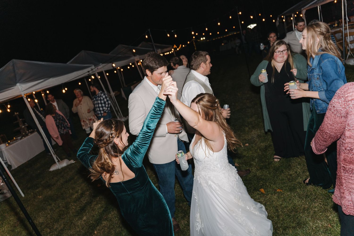 Guests celebrate under a tent at an outdoor evening wedding reception with string lights overhead.