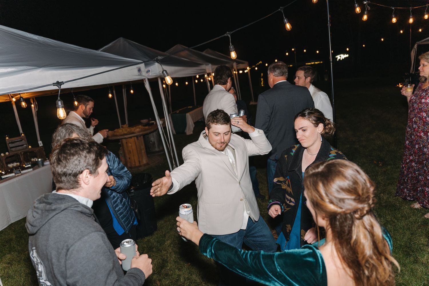 A group of people dancing and socializing under string lights at an outdoor tent party.