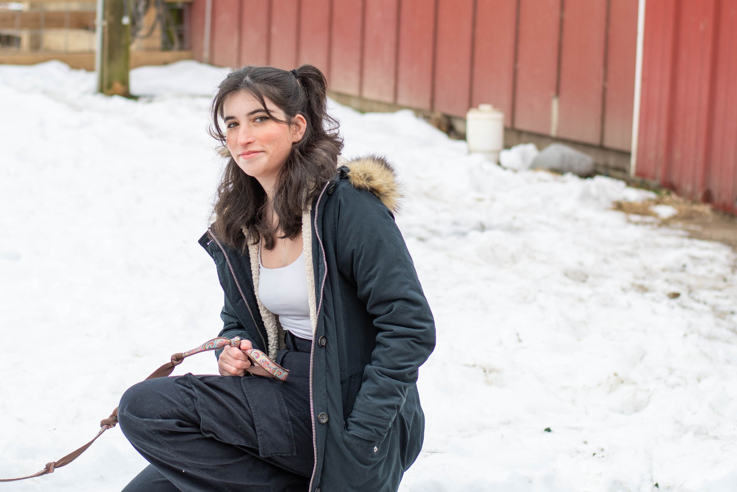 A woman in a winter coat sits outdoors in the snow near a red barn.