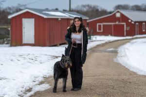 A person and their large black dog stand on a path with red barns and snow in the background.