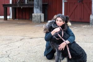 A tender moment between a person and their black dog outside a red barn.
