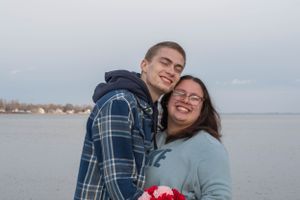 A couple shares a tender moment by a waterfront on an overcast day while holding red flowers.