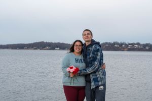 A couple poses together by a lakefront with red flowers and houses visible in the background.