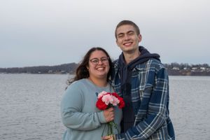 A joyful couple poses together by a lake holding red flowers on an overcast day.
