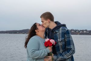A romantic moment captured between a couple sharing a kiss by the water while holding red flowers.