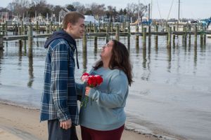 A couple shares a romantic moment at a marina dock with one holding red roses.