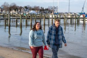 A couple walks along a marina beach with boats and docks in the background.