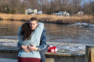 A couple embraces on a dock with waterfront homes and melting ice visible in the background.