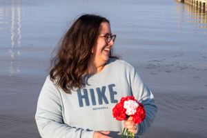 Person in a light blue HIKE sweatshirt smiles while holding red flowers by the water.