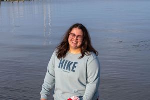 Person wearing glasses and a HIKE sweatshirt smiles while holding flowers near the water.