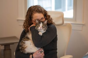 Person holds a calico cat while sitting in a white chair by a window.