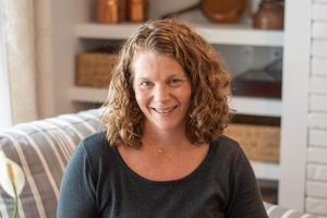 A woman with curly brown hair smiles warmly while sitting in a casual home setting with shelves in the background.