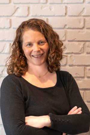 Portrait of a person in black sweater standing against white brick wall with confident crossed arms pose.