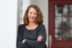 Professional portrait of someone in dark sweater with crossed arms against white exterior wall.