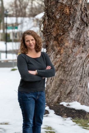 Winter outdoor portrait by large tree trunk with snow on ground wearing jeans and dark top.