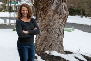 Person in casual attire poses confidently next to large tree trunk in snowy outdoor setting.