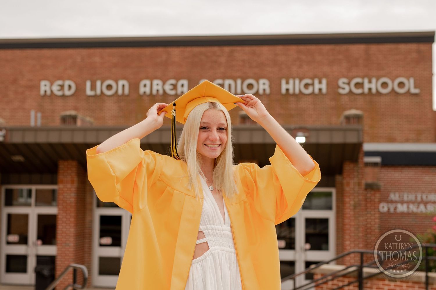 Graduate in yellow gown adjusts cap while standing outside high school entrance.