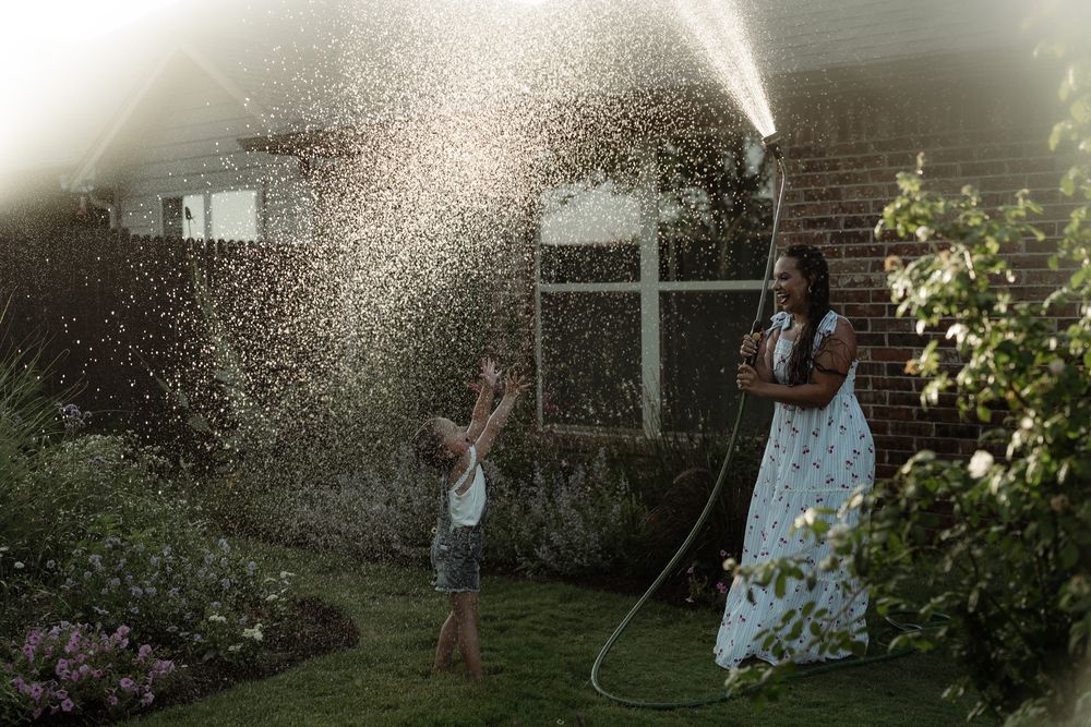 Woman sprays garden hose as child plays in water mist in lush backyard on summer evening.