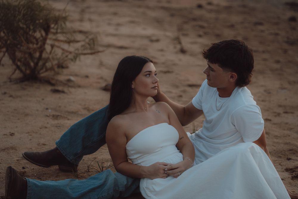Two brides in white and blue gowns share a tender moment sitting together on sandy desert ground.