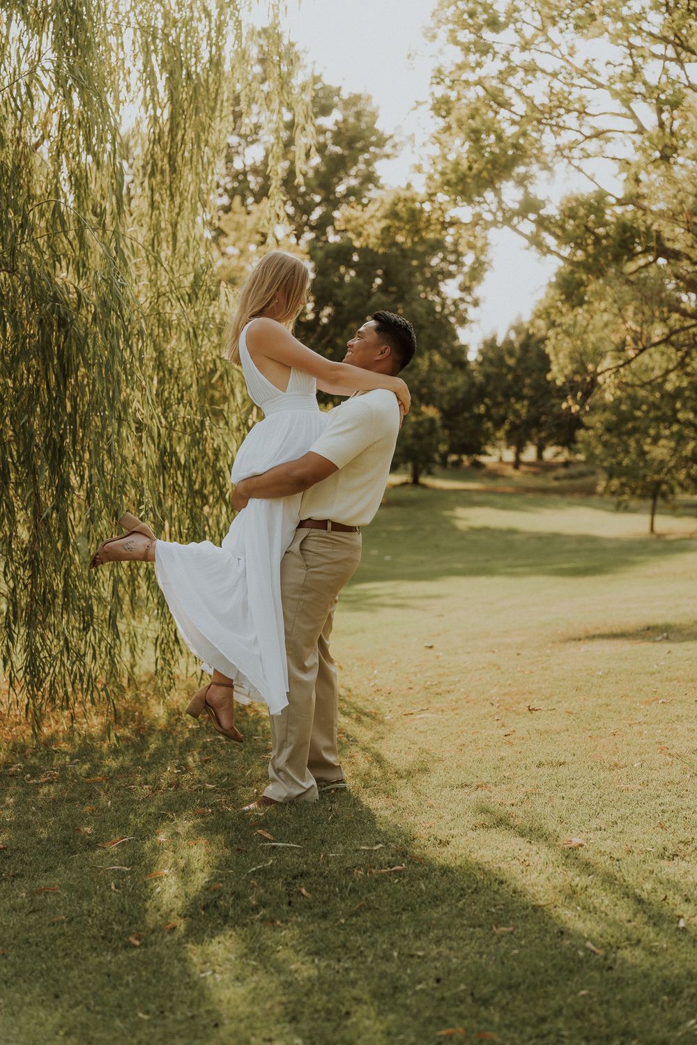 A man lifts a woman in a white dress near a weeping willow tree in a romantic outdoor setting.