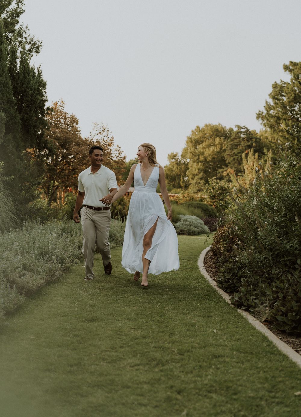 A couple walks hand-in-hand through a lush garden path, the woman in a flowing light blue dress, both smiling joyfully.