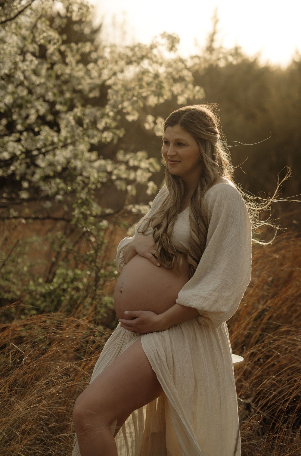 A pregnant woman in a flowing white dress poses in a field at golden hour with blossoming trees in the background.