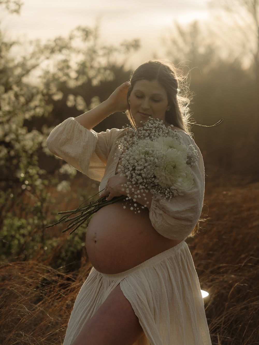 Ethereal maternity photo in white dress holding baby's breath flowers at golden hour in nature.