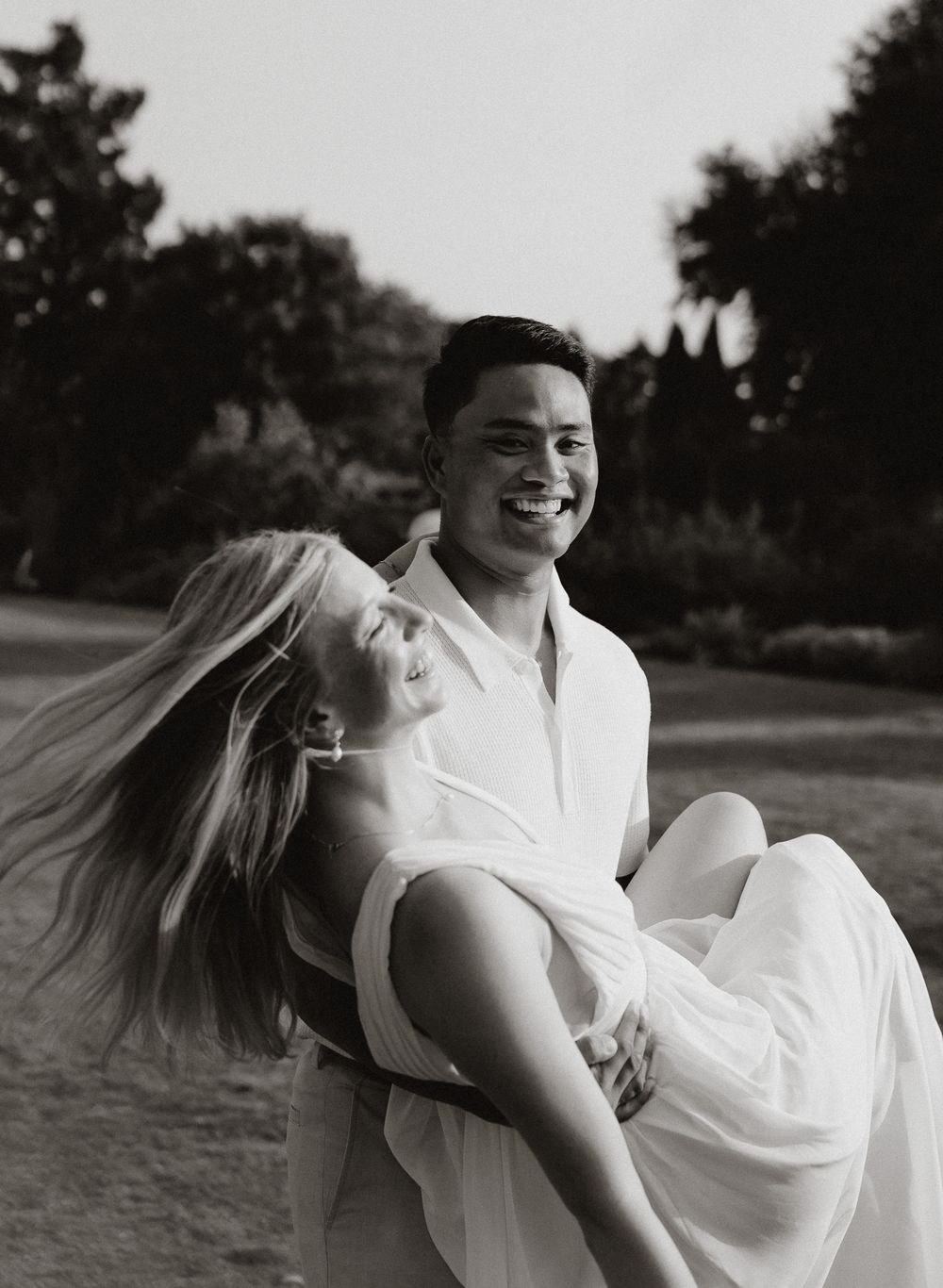Black and white photo of a joyful couple, man lifting laughing woman in a park setting.