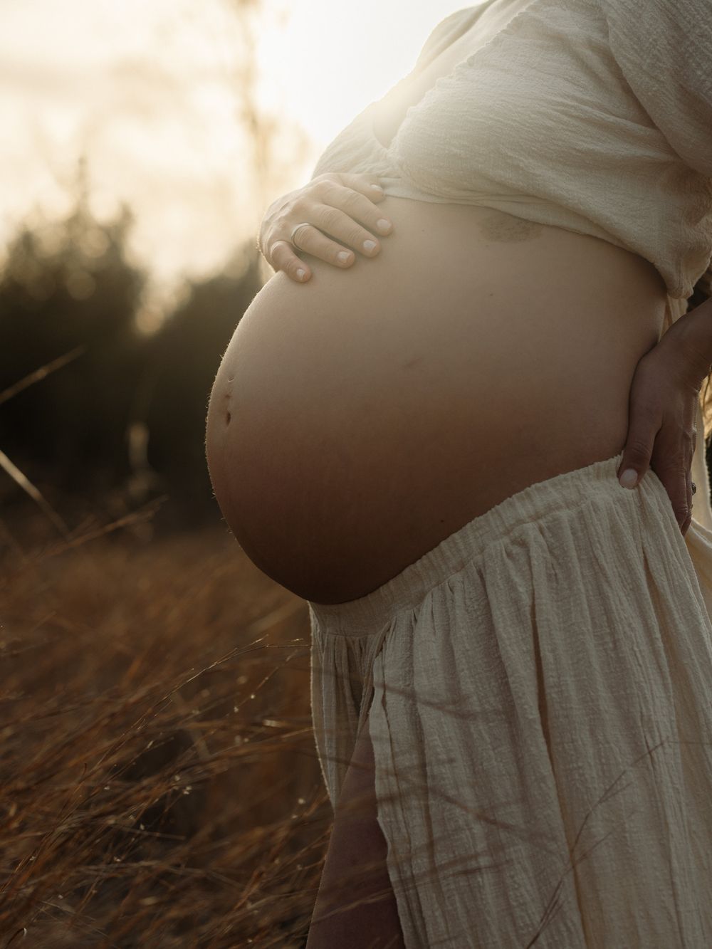 A pregnant belly silhouetted against golden sunlight while wearing a flowing white dress in a field at sunset.
