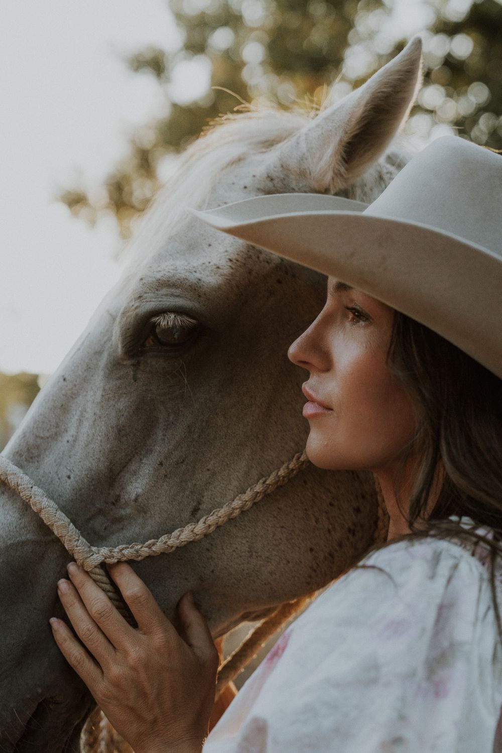 Woman in white cowboy hat tenderly resting her head against a white horse, sharing a peaceful moment outdoors.