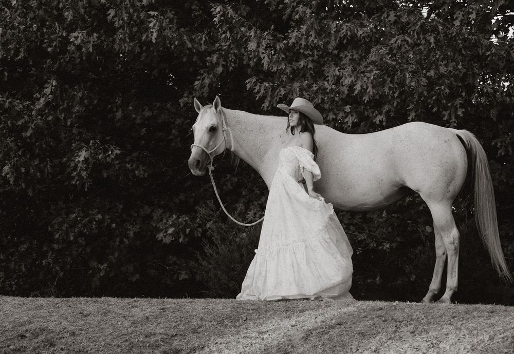 Woman in white dress stands beside a white horse in a black and white outdoor photograph.