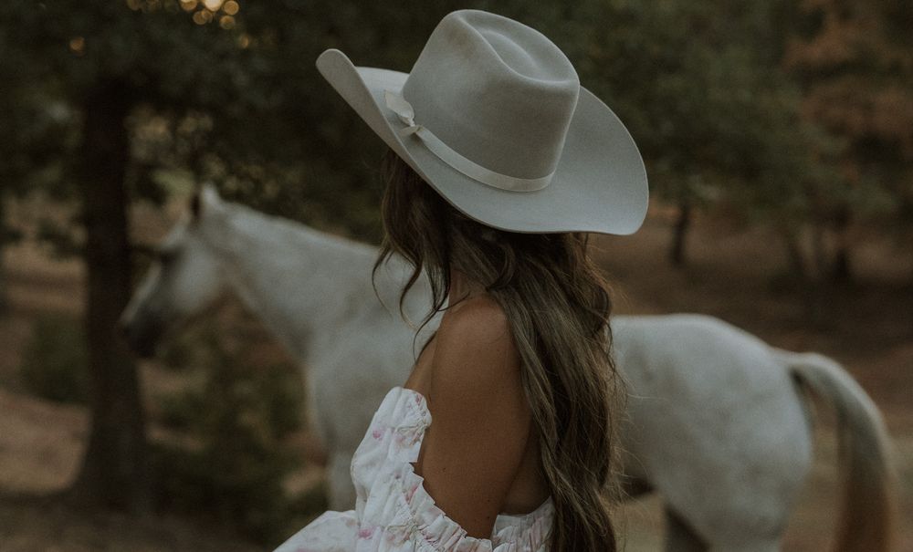 Woman in white dress and cowboy hat standing near a white horse outdoors in a wooded setting.