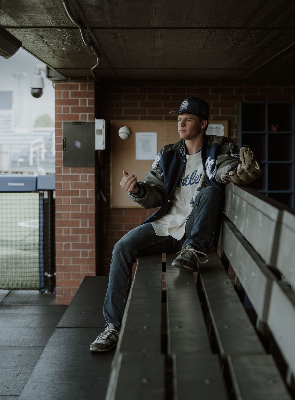 Young man in baseball cap and jersey sits in a dugout bench holding a glove at a baseball stadium.