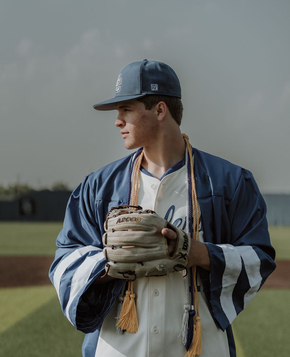 Young baseball player in navy uniform and cap holds glove on field, wearing a blue warm-up jacket with gold tassel.