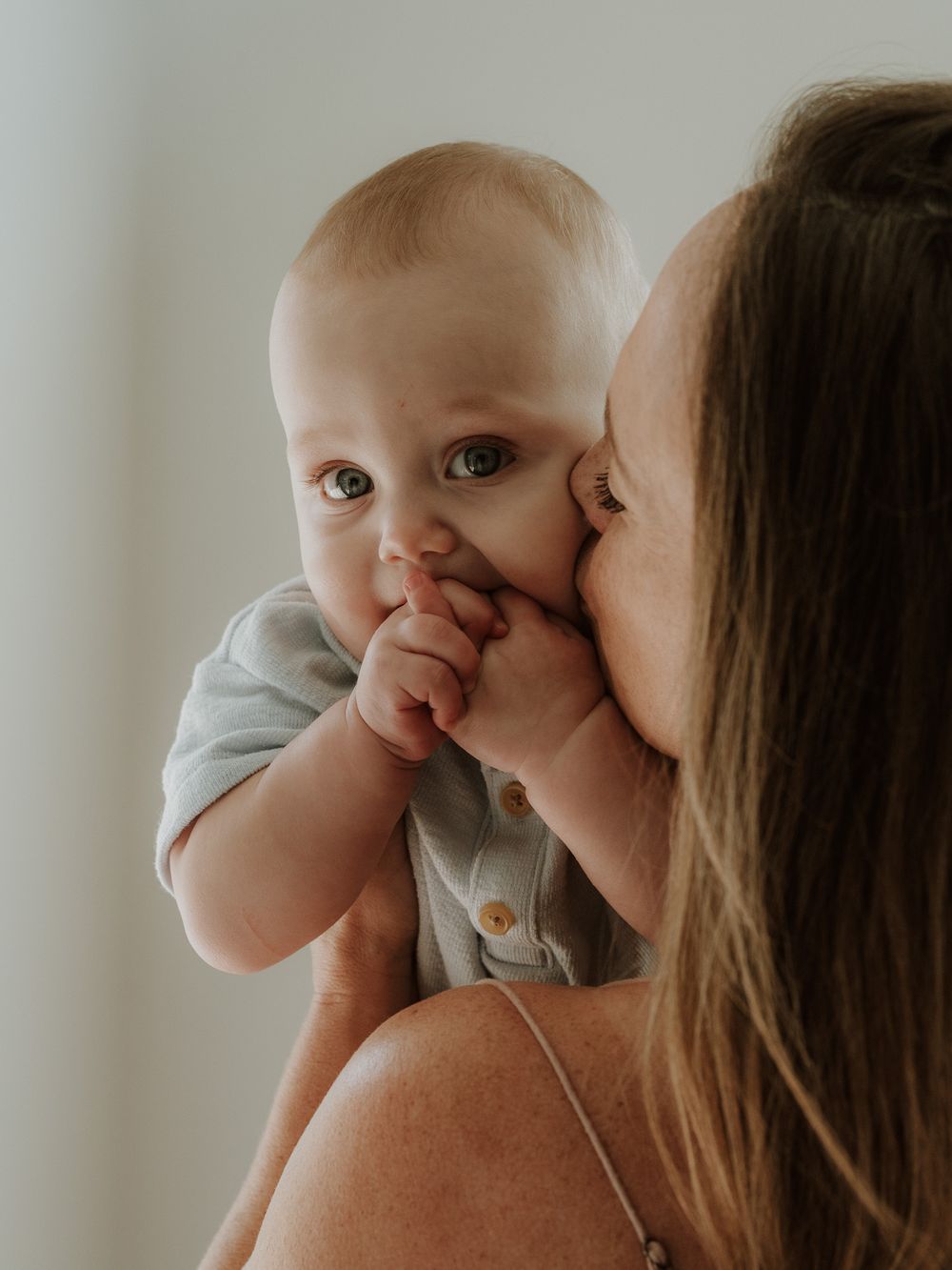 A mother tenderly kisses her smiling baby in a light blue onesie against a soft white background.