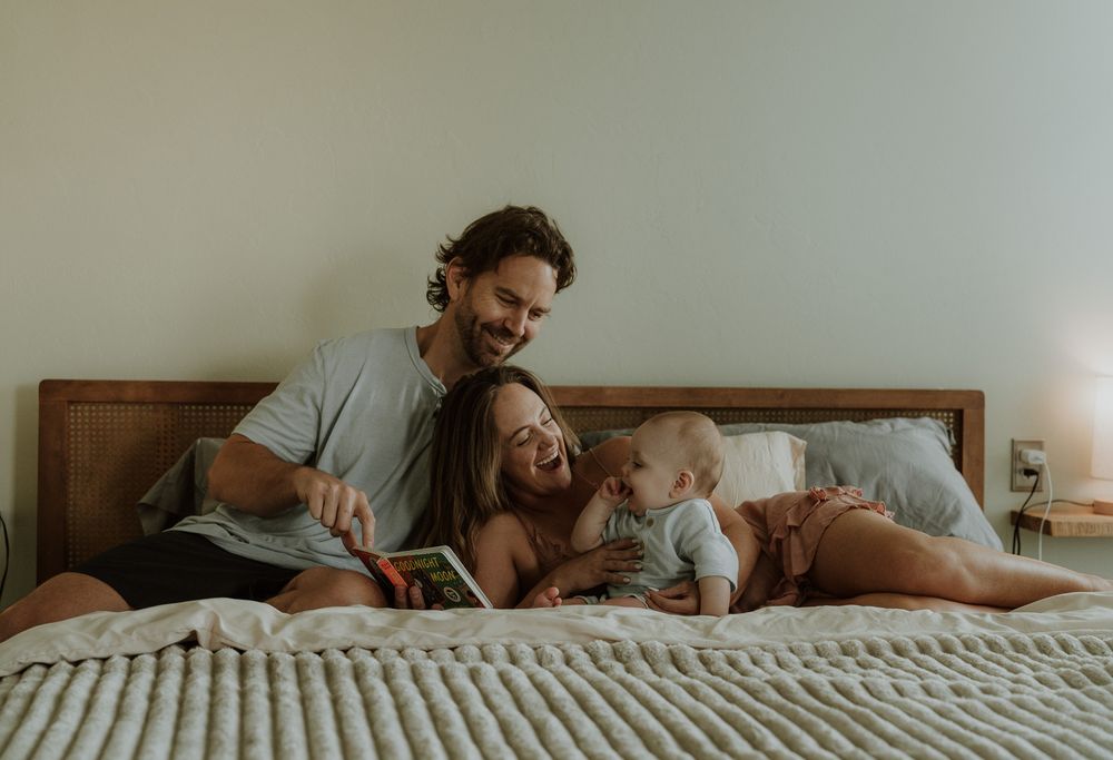 Happy family with parents and baby relaxing on bed, reading a colorful book together in a bright bedroom.