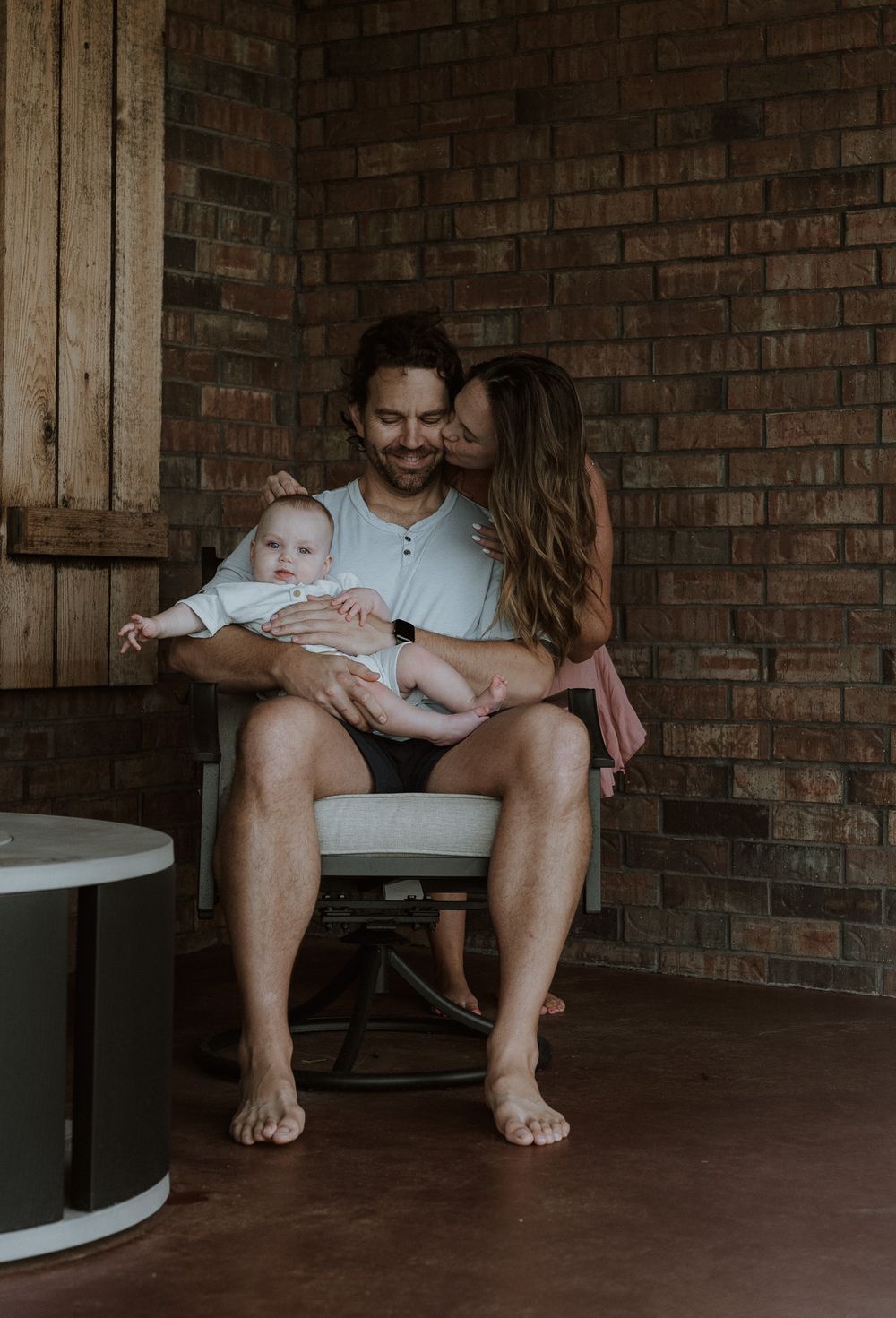 A smiling man holds a baby in a chair while a woman kisses his cheek near a brick wall.