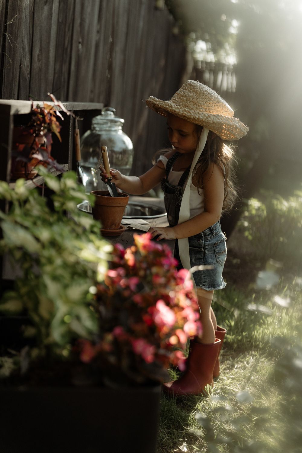 Young girl in straw hat, denim overalls, and red boots tending to potted plants in a sunny garden.