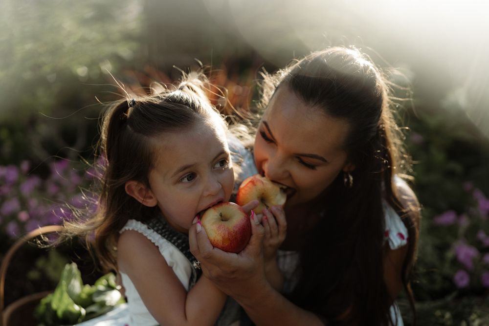 A mother and young daughter share an apple outdoors in soft sunlight, surrounded by blooming flowers.