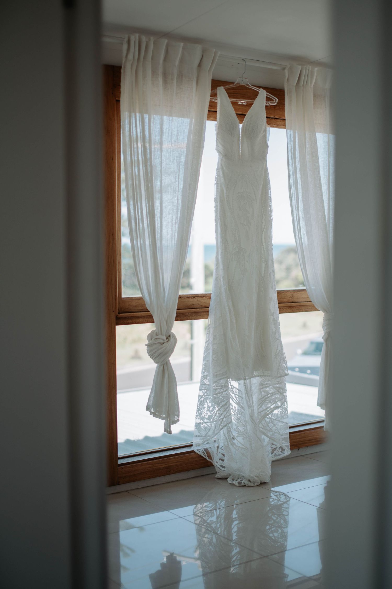 White lace wedding dress hanging in window with sheer curtains and natural light.