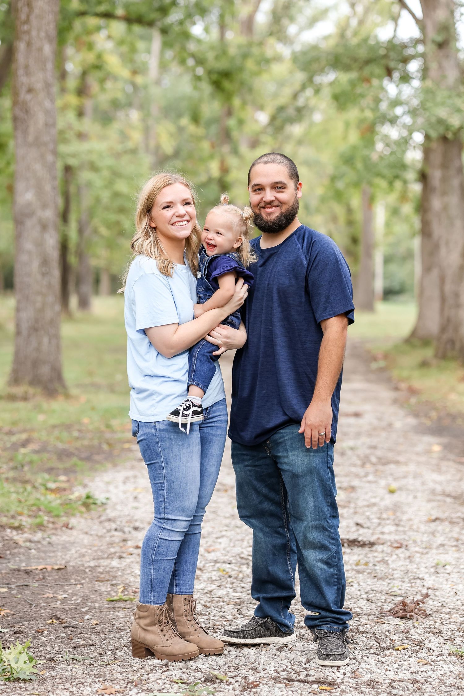 A family stands together on a nature path surrounded by tall trees and greenery in casual outdoor attire.