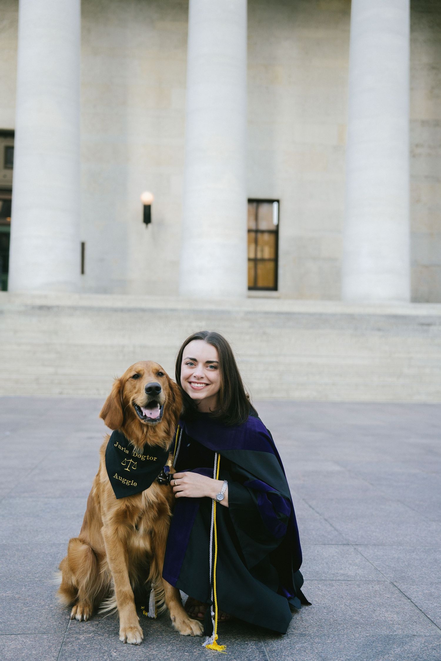 Graduate in cap and gown poses with friendly golden retriever wearing a bandana outside a columned building.