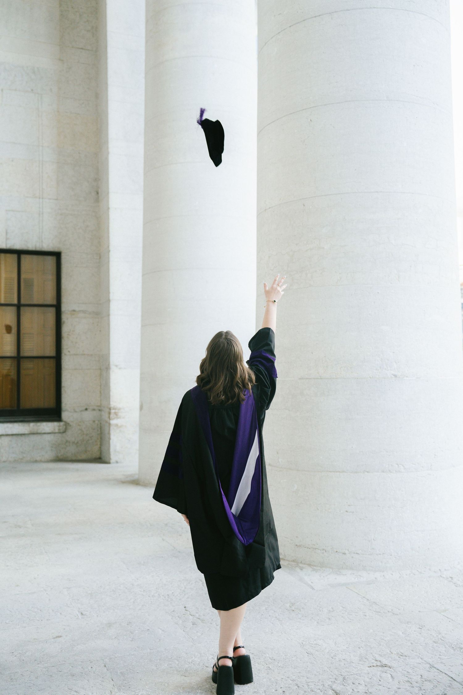 Graduate tosses cap in celebration between white pillars during graduation ceremony.