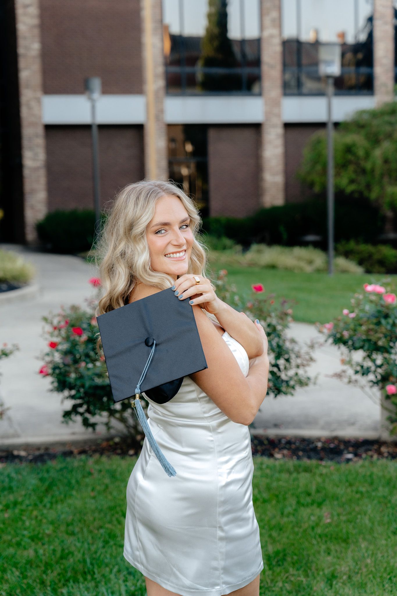 Graduate in white satin dress poses with graduation cap near blooming rose garden.