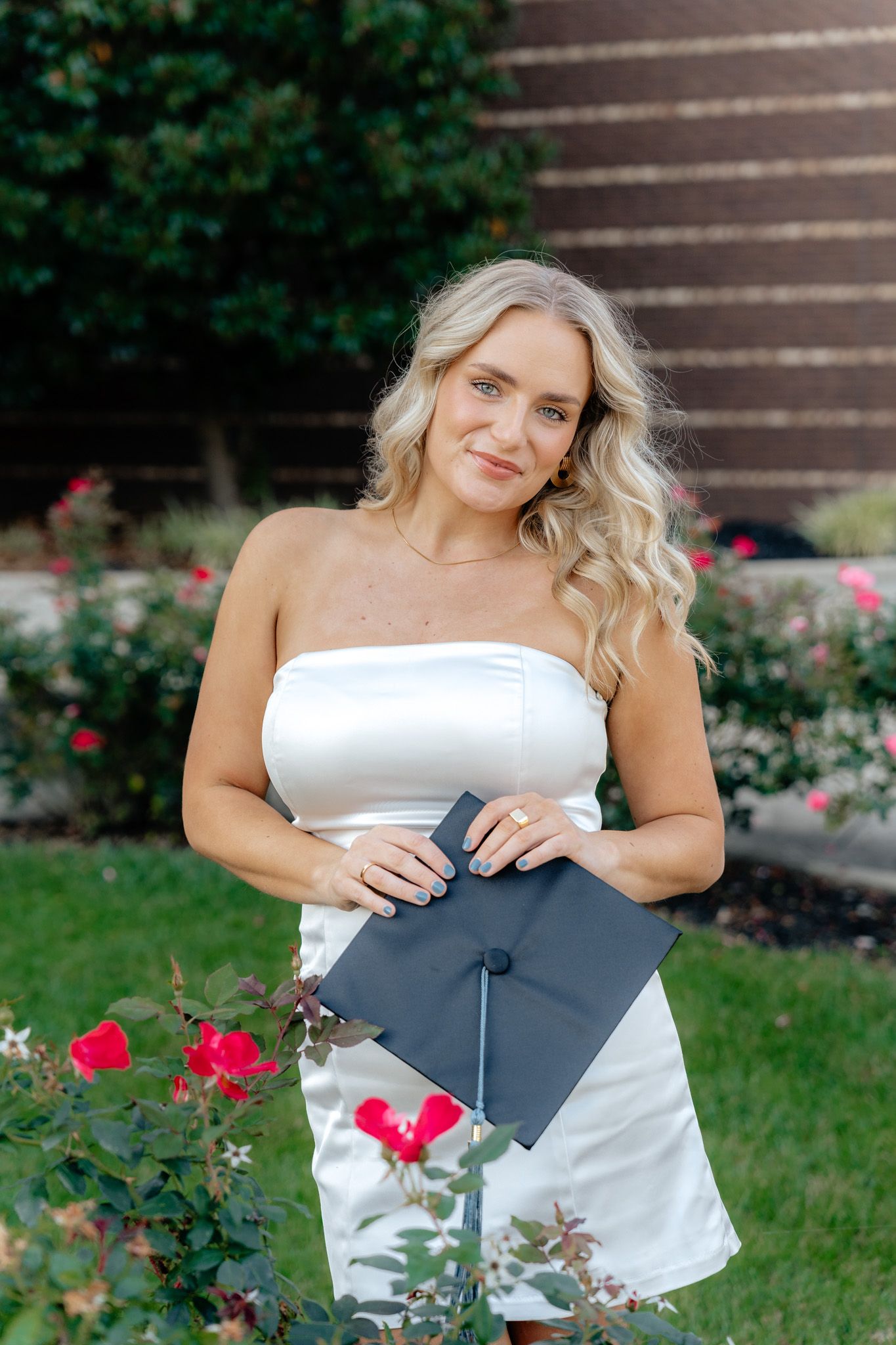 Smiling graduate in strapless white dress stands among pink roses with graduation cap.
