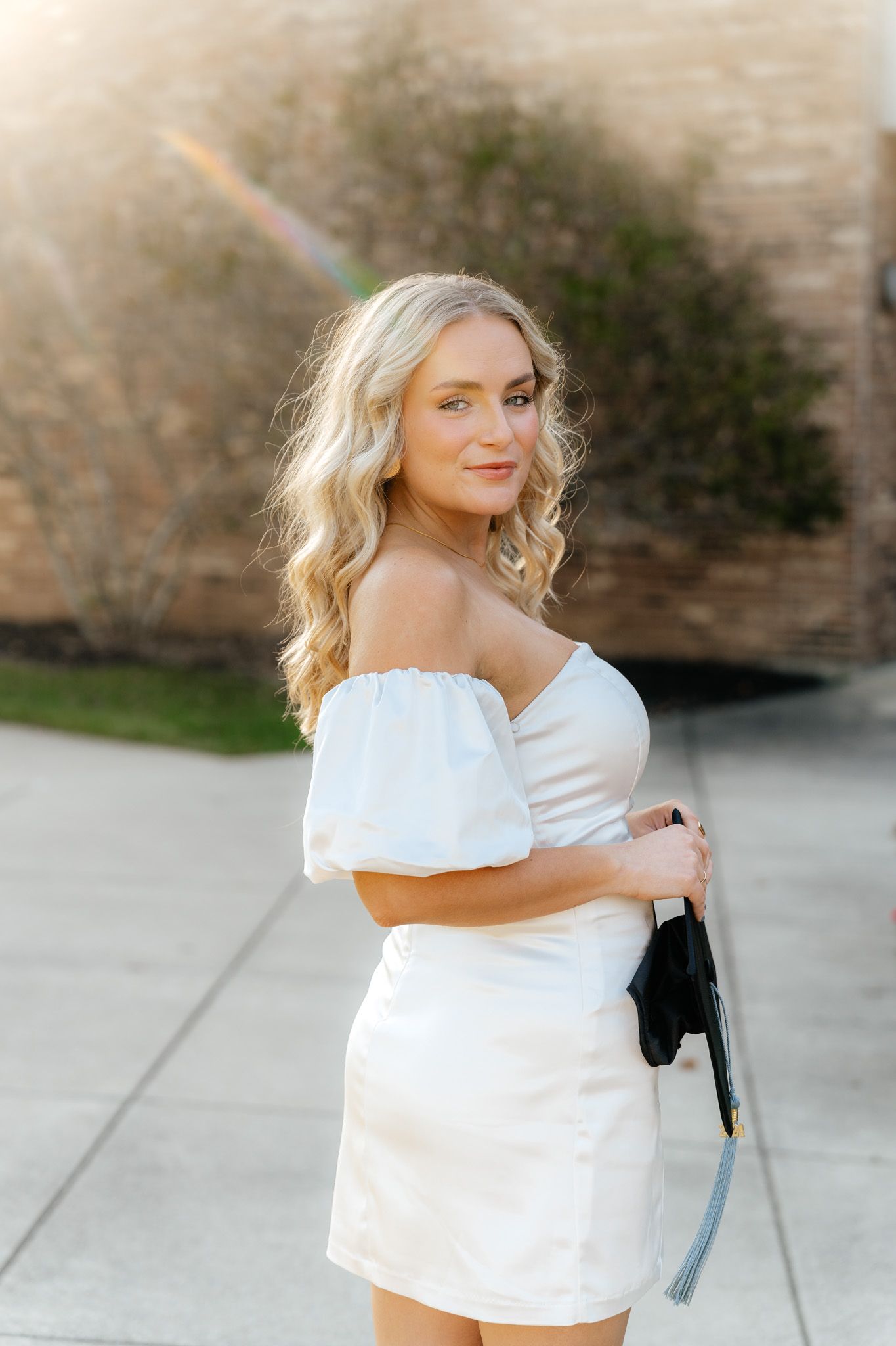Graduate in off-shoulder white dress poses outside building during sunset.